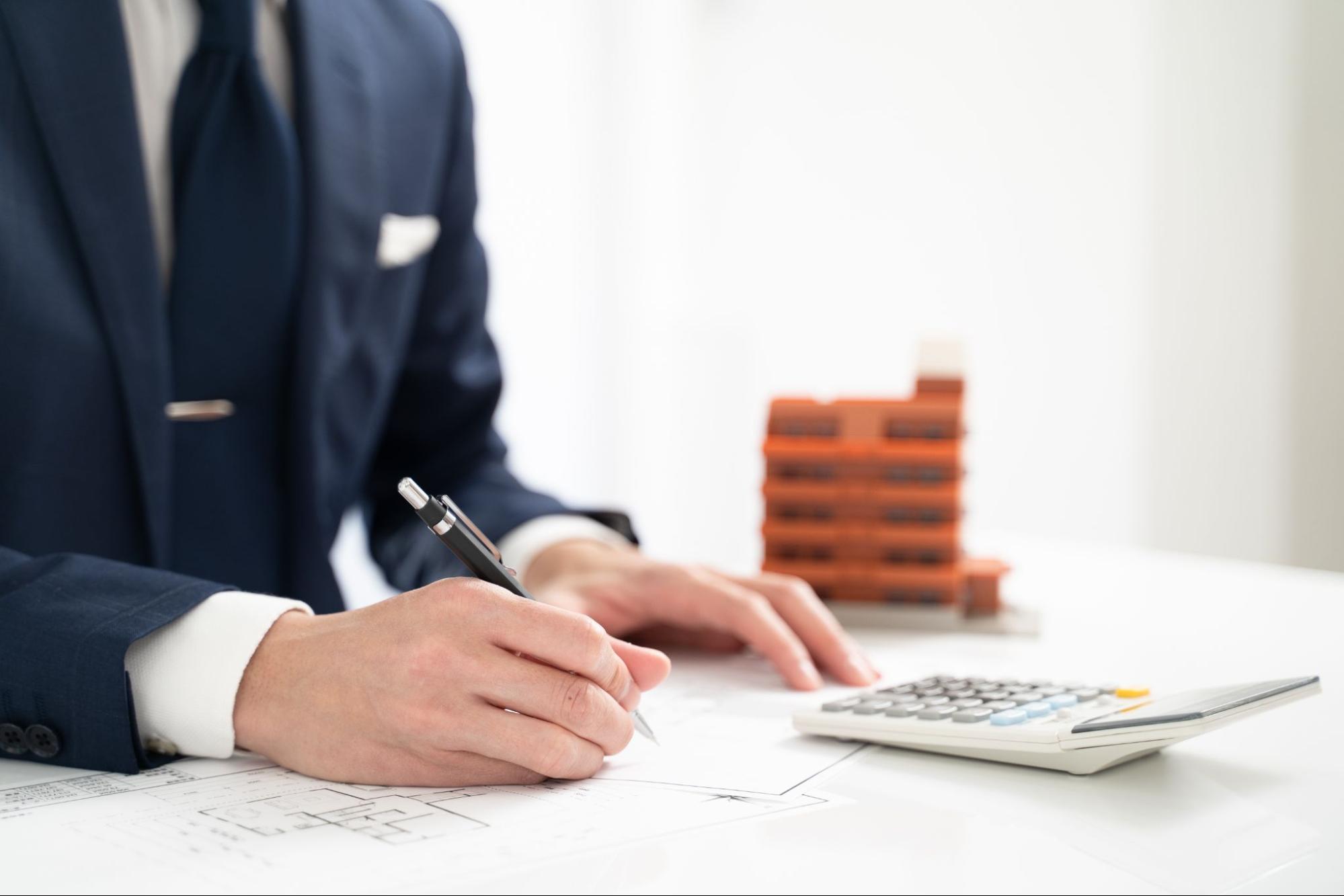 image9 | 不動産の売却・査定のお役立ちメディア不動産売却マスター Businessperson in a navy suit writes on documents at a desk, with a calculator and blueprints nearby.